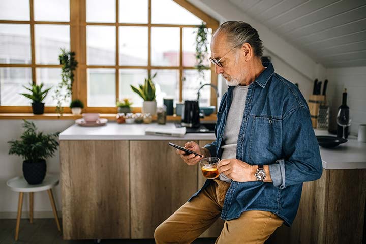 Mature man in kitchen looking at phone holding coffee.